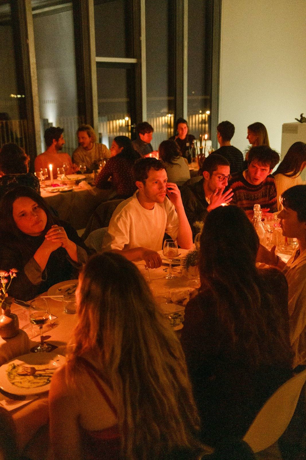 Guests seated at long candlelit tables during a supper club, the windows behind them fading to dusk.